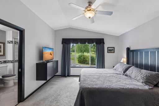 Vaulted ceilings enhance the upper level primary bedroom, complemented by carpeted flooring, a large window, ceiling fan with light, and a walk-in closet with custom built-in storage.