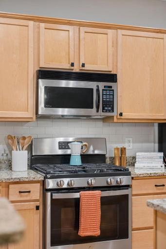 Stainless steel gas range and microwave surrounded by warm wood cabinetry and tiled backsplash.