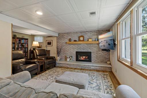 The lower-level family room offers a cozy gathering space centered around a brick fireplace with a raised hearth and wood mantel. Large windows bring in natural light while providing views of the backyard.