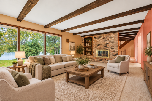 Spacious living room featuring exposed beams, brick fireplace, and large windows showcasing beautiful water views. Virtually Staged