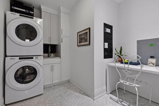Carrara marble flooring, custom JW Woodcraft cabinetry and a black herringbone backsplash elevate the laundry room. A Trinity utility sink with Kohler touchless faucet and custom built-in desk add exceptional function.