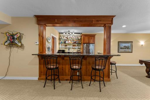 The spacious recreation area includes a nearby wet bar with high-top seating, a full-size refrigerator, ample cabinetry and counter space, a cabinet-front dishwasher, and additional shelving.