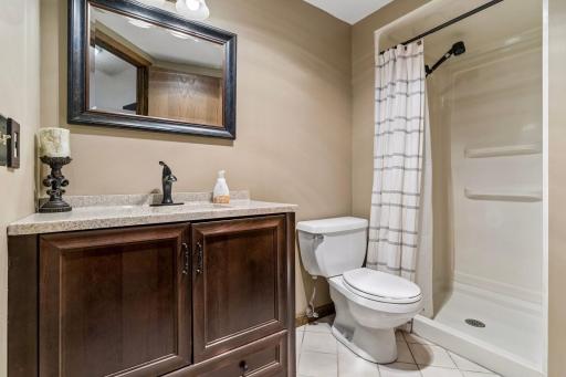 Convenient lower level bathroom featuring a dark wood vanity with a stone-look top, framed mirror, tile flooring, and walk-in shower stall.