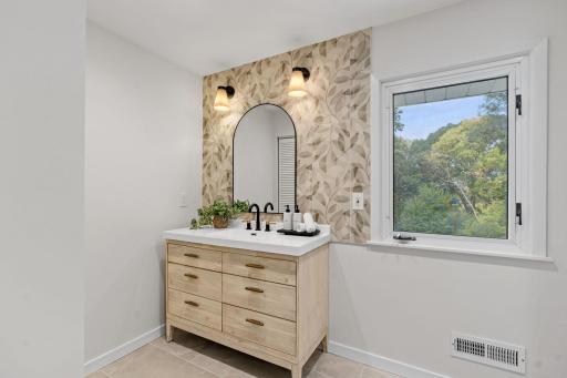 The beautifully updated bathroom features a furniture-style vanity, contemporary fixtures, botanical tiled back splash, and peaceful views of the surrounding trees.