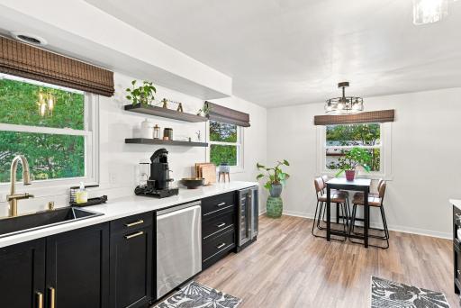 Striking BRAND-NEW eat-in kitchen featuring sleek stainless steel appliances, luxurious quartz countertops, striking black cabinetry, elegant gold hardware, contemporary light fixture, wine fridge, newer flooring, windows, & stylish window coverings.