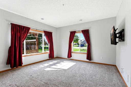 Spacious living room featuring plush carpeting and two large windows that fill the space with abundant natural light. The window treatments and TV wall mount will remain with the home. TV will be removed.