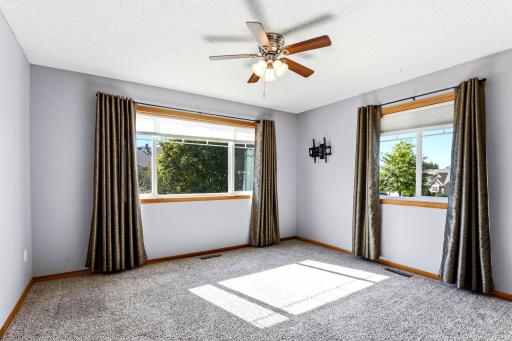 Bright upper-level bedroom with plush carpeting, ceiling fan, and two large windows. Window treatments and TV wall mount are included.
