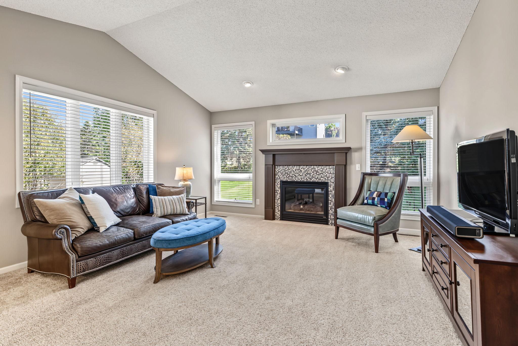 Inviting main-level living room featuring a cozy gas fireplace with a beautiful tile surround, flanked by windows that fill the space with natural light. The vaulted ceiling adds to the spacious, airy feel.