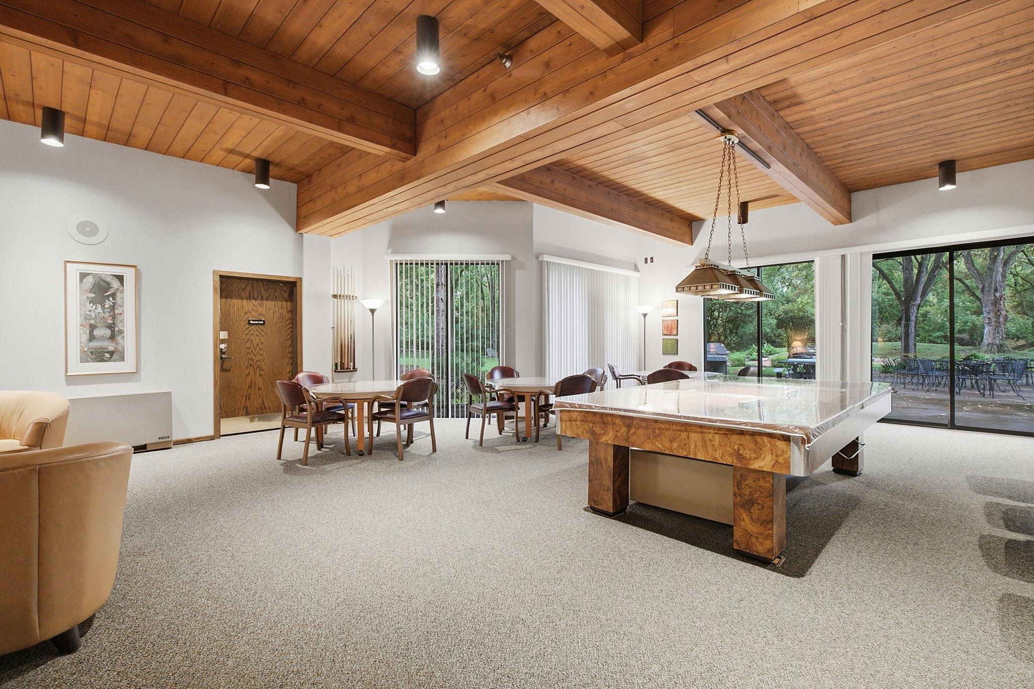 Community Room - Spacious community room with a billiards table, multiple seating areas, distinctive wood-beamed ceiling, and large windows overlooking the outdoor grounds.