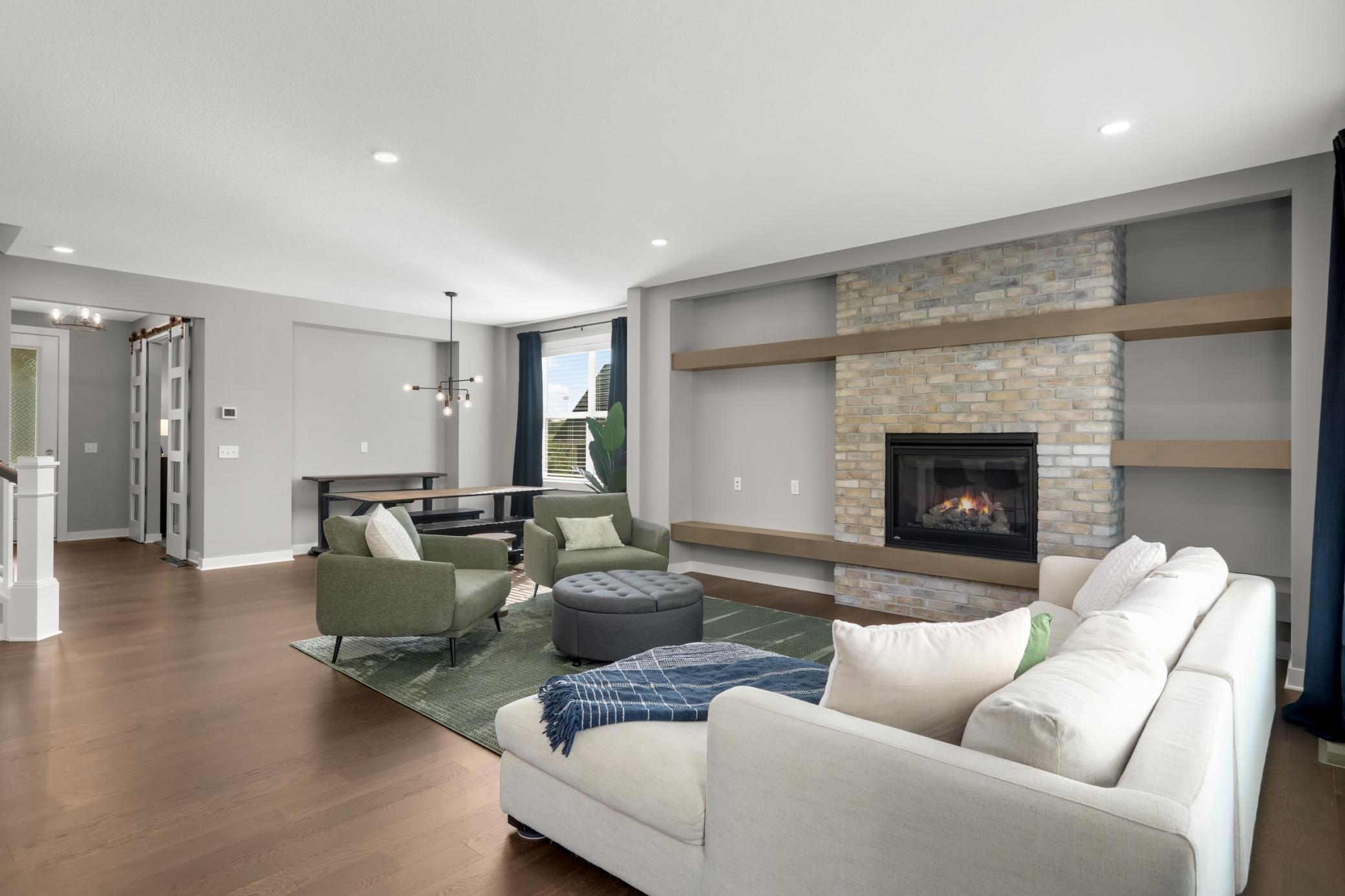 Spacious living room highlighted by a statement fireplace, brick surround, custom floating shelves, and hardwood floors.