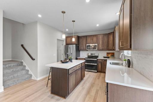 A bright, spacious kitchen featuring quartz countertops, rich cabinetry, stainless steel appliances, tile backsplash, and a large center island with seating. The open layout flows seamlessly into the dining and living spaces.