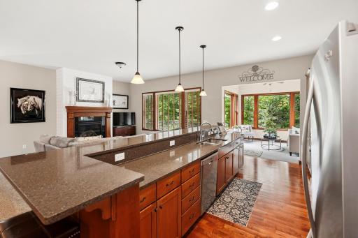 Warm, inviting kitchen with wooden cabinets and stainless steel appliances, complemented by a cozy living area with a granite countertop and pendant lighting. Large windows flood the space with natural light, creating a welcoming atmosphere.