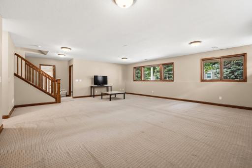 Cozy living space with warm lighting and neutral tones, featuring a carpeted floor, wooden staircase, and large windows offering a serene view of the outdoors. The room is well-lit with ceiling lights, creating a comfortable and inviting atmosphere.