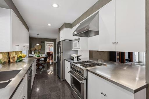 Kitchen - Bright workspace features a double sink beneath a wide window, stainless steel counters, dishwasher, white cabinetry, gas range, and tile flooring.