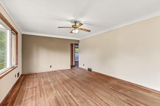Living room view highlighting the wide picture window, hardwood floors, ceiling fan, and flexible furniture space.
