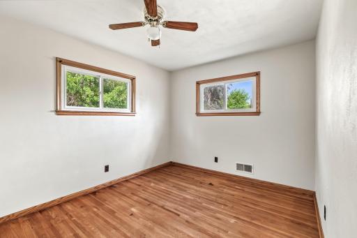 Bright main-level bedroom with hardwood floors, two windows, ceiling fan, and neutral finishes.