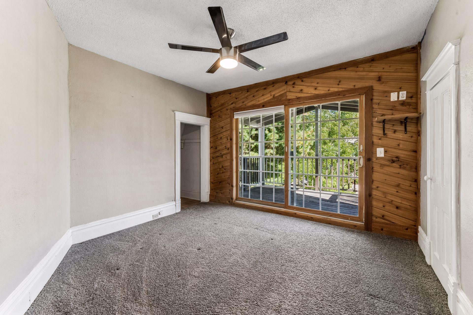The inviting primary bedroom features a contemporary ceiling fan, warm wood accent wall, and sliding glass doors leading to a private covered deck overlooking the front of the home.
