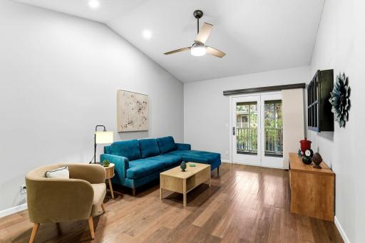 Bright, spacious living room featuring vaulted ceilings, recessed lighting, a ceiling fan, wood flooring, and French doors opening to a private balcony.