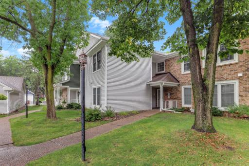 Exterior view of 10966 Quebec Ave S featuring brick and vinyl siding, mature shade trees, lush green lawn, landscaped gardens, and winding sidewalks in a peaceful setting.