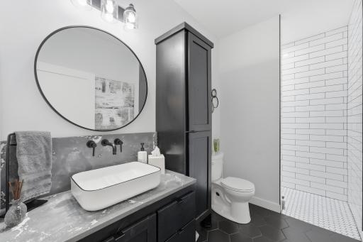 Modern bath features dramatic dark cabinetry, generous storage, vessel sink, wall-mounted fixtures, Kohler toilet, bluetooth fan, heated tiled flooring, and a beautifully finished walk-in shower. Conveniently located next to beds & front entry.