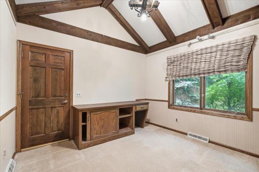 Versatile main-level office or flex space features a vaulted beamed ceiling, built-in cabinetry and workspace, and a large window overlooking the wooded backyard.