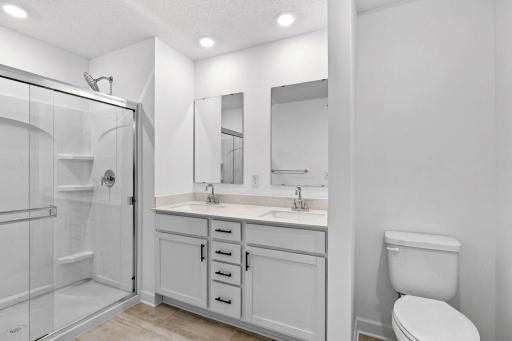 Featuring a glass-enclosed shower with built-in shelving and glass mirrors set atop dual quartz sinks and vanity-cabinetry, the en-suite bathroom helps streamline morning routines