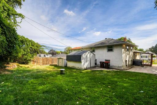 Rear exterior view showcasing the covered deck, expansive driveway, yard, and one of two large storage sheds.