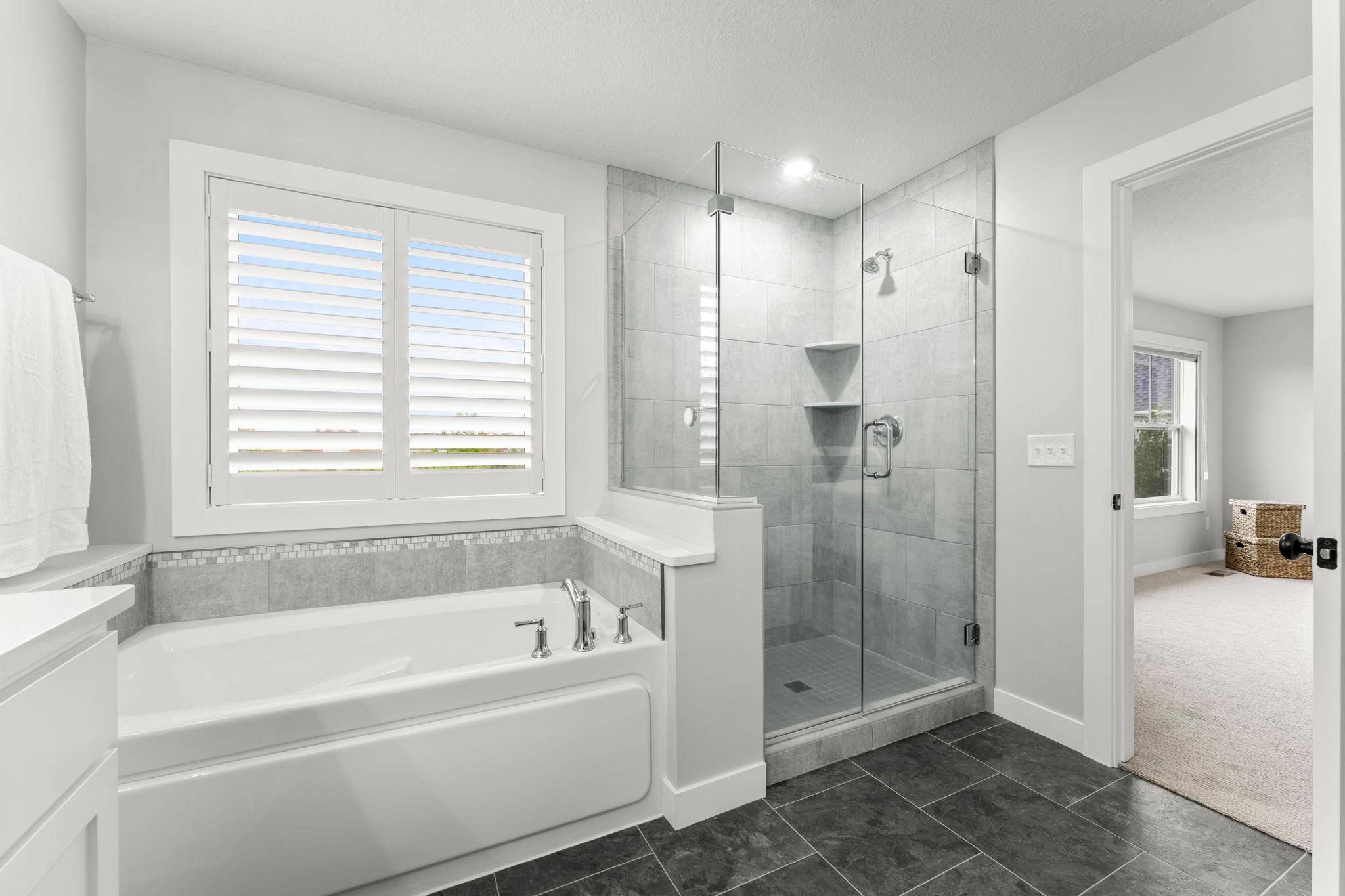 This view of the primary full bathroom highlights the glass shower, soaking tub and the ceramic tile surround, and the large window.
