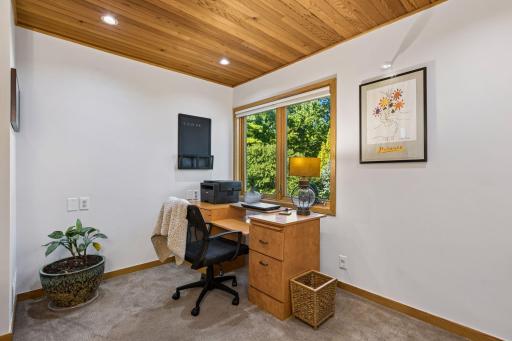 Bright private office with built-in desk space, large windows overlooking the mature greenery, wood-plank ceiling, and ample room for working from home.