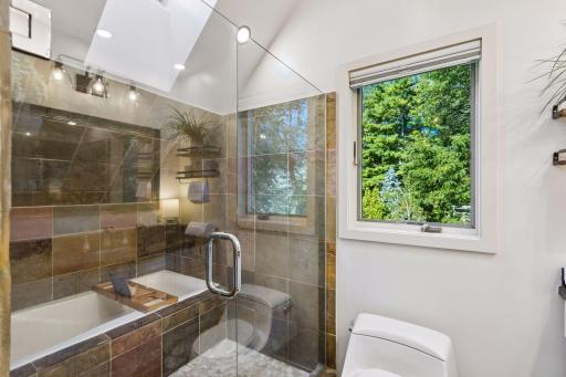 Beautifully finished en-suite bathroom includes a tiled tub and shower surround, glass shower door, modern fixtures, and a large window bringing in natural light and leafy views.