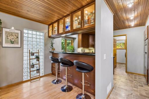 Warm and inviting kitchen featuring rich wood cabinetry, glass-front display cabinets, a wood-plank ceiling, breakfast bar seating, and abundant natural light.