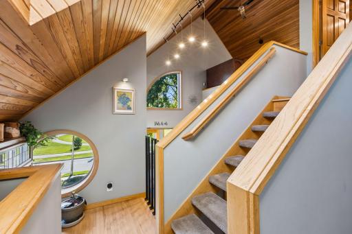 Dramatic open staircase connects the levels beneath a warm wood-paneled vaulted ceiling, with custom railings, multiple arched windows, and hardwood flooring adding architectural character.