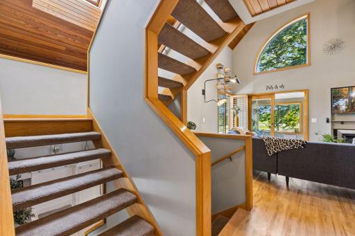 Custom wood staircase with carpeted treads and warm wood railings opens to the main living area, showcasing vaulted ceilings, hardwood floors, abundant natural light, and distinctive arched windows.