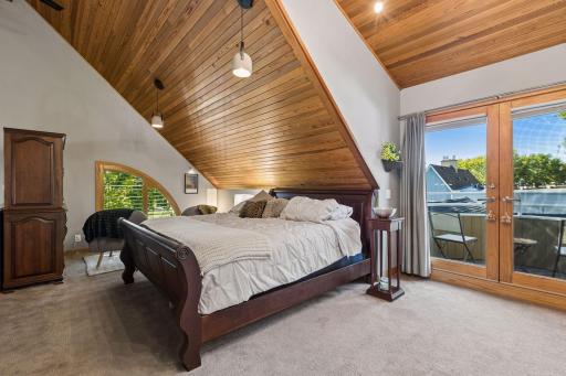 Spacious primary bedroom features a dramatic vaulted wood-paneled ceiling, abundant natural light, plush carpeting, custom built-ins, and French doors opening to a private balcony.