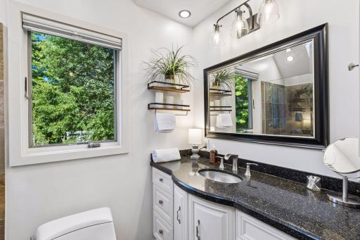 The en-suite bathroom features white cabinetry, contemporary lighting, large framed mirror, and a bright window overlooking mature greenery.