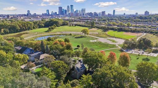 Expansive aerial perspective overlooking lush parkland, walking paths, recreational fields, and the Minneapolis skyline beyond.