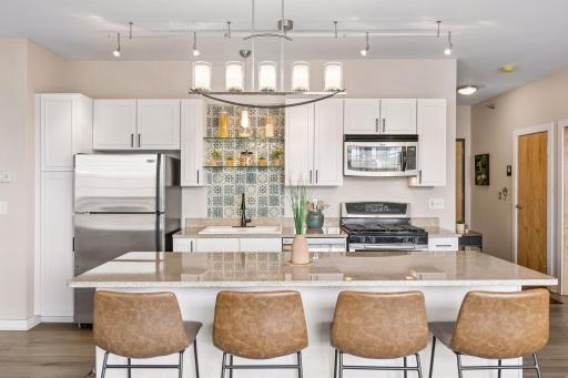 This well-appointed kitchen showcases abundant cabinetry, stone countertops, stainless steel appliances, large island, and glass shelving against a decorative tile backsplash.