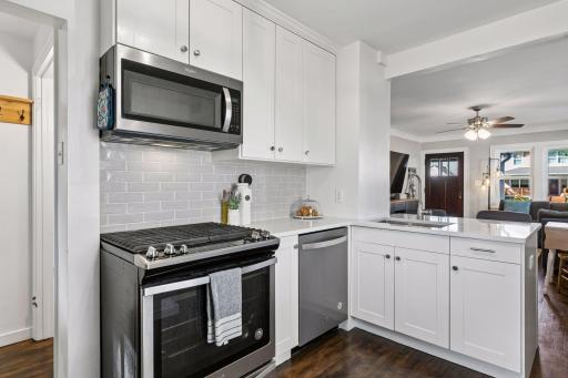 Modern kitchen featuring a gas range, stone countertops, farmhouse sink, white cabinetry, stainless-steel appliances, tiled backsplash and breakfast bar overlooking the main living area.