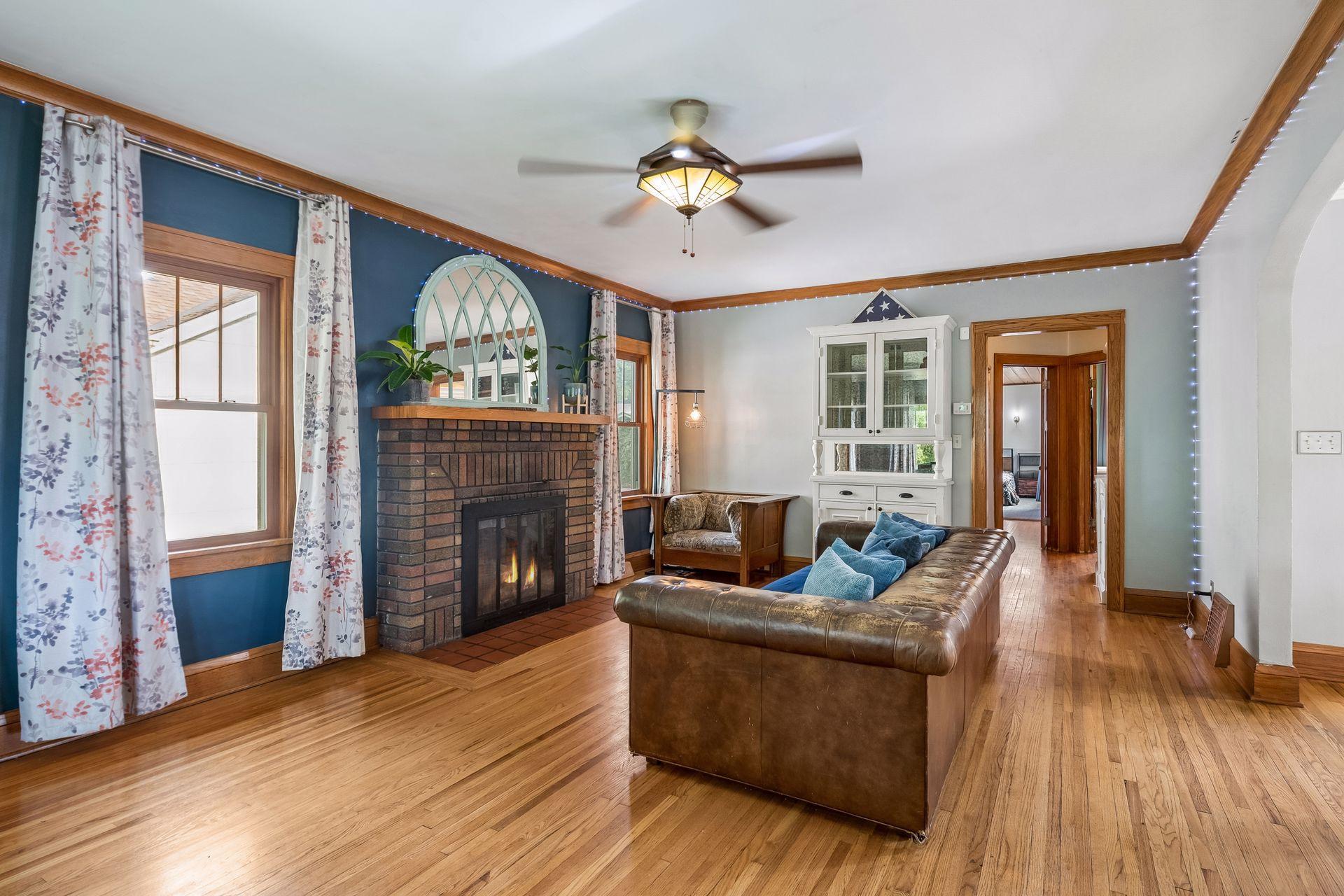 The heart of the home is the cozy living room, anchored by a classic brick fireplace with a wood mantel. A deep blue accent wall and windows on both sides make it warm and bright at once.