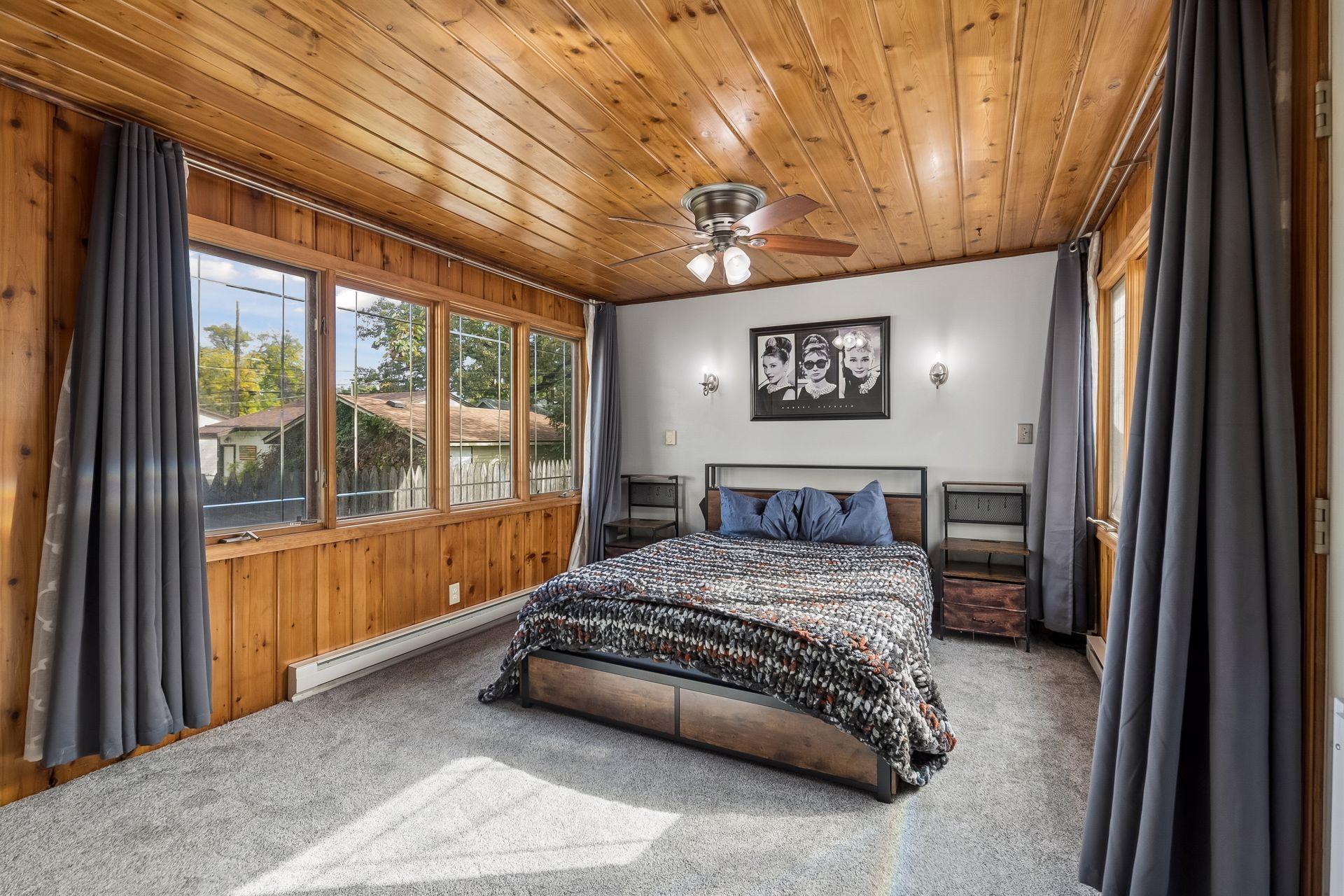 The primary bedroom is full of charm, with knotty wood paneling on the walls and ceiling, a ceiling fan, and a wall of windows. New carpet with plush padding feels great underfoot.