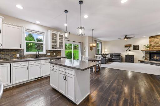 Bright, open-concept kitchen features a large center island, abundant cabinetry, and pendant lighting overlooking the dining and living areas. Enjoy seamless indoor-outdoor living with backyard access and a stunning stone fireplace as the focal point