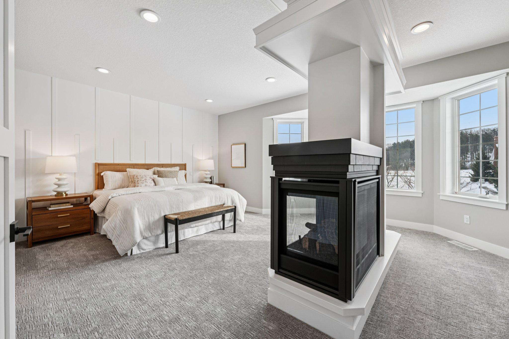 Spacious primary suite showcasing a dramatic two-sided fireplace, soft neutral tones, and oversized windows that fill the room with natural light.