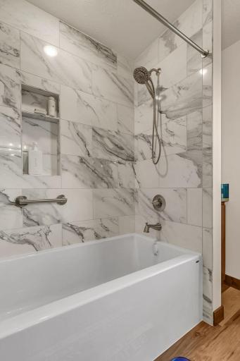 Pristine bathroom boasts an oversized soaking tub framed by stunning, floor-to-ceiling marble-patterned tile work with dramatic veining. High-end hardware, a sleek built-in shower niche and matching safety grab bar.