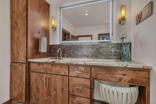 Custom designed primary vanity, showcases beautiful grain patterns, built-in hamper, premium stone countertops, earthy mosaic backsplash, integrated lighting with modern sconces, large wide-profile lighted mirror.