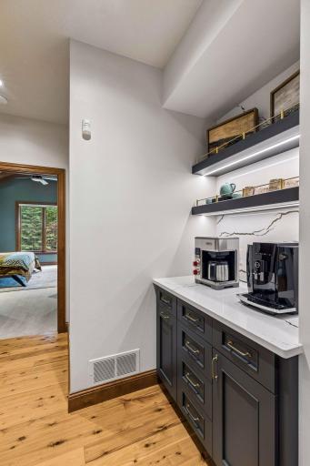 Coffee bar with quartz counters, illuminated floating shelves, and brass-accented cabinetry, conveniently located between the kitchen and the primary suite
