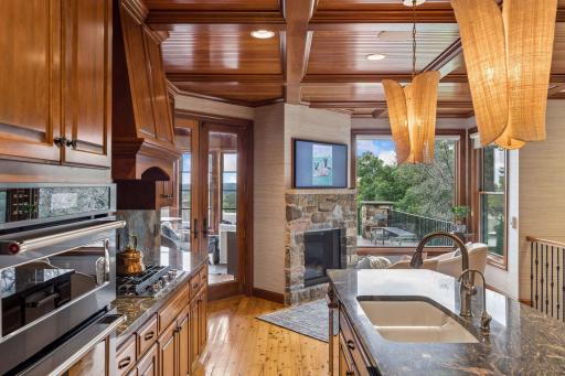 Gourmet kitchen with a coffered beadboard ceiling, custom cherry cabinetry, slate backsplash, and granite counters, opening to a cozy keeping room with a stone fireplace and porch access
