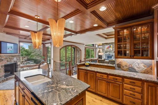 Gourmet kitchen with a coffered beadboard ceiling, custom cherry cabinetry, slate backsplash, and granite counters, opening to a cozy keeping room with a stone fireplace and porch access