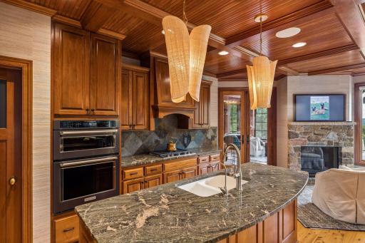 Gourmet kitchen with a coffered beadboard ceiling, custom cherry cabinetry, slate backsplash, and granite counters, opening to a cozy keeping room with a stone fireplace and porch access