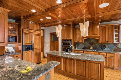 Gourmet kitchen with a coffered beadboard ceiling, custom cherry cabinetry, slate backsplash, and granite counters, opening to a cozy keeping room with a stone fireplace and porch access