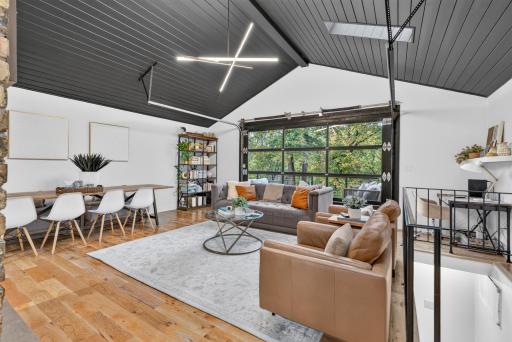 Another view of the open living room highlighting the vaulted ceiling, hardwood floors, dining area and glass garage door overlooking the backyard and access to the lower level.